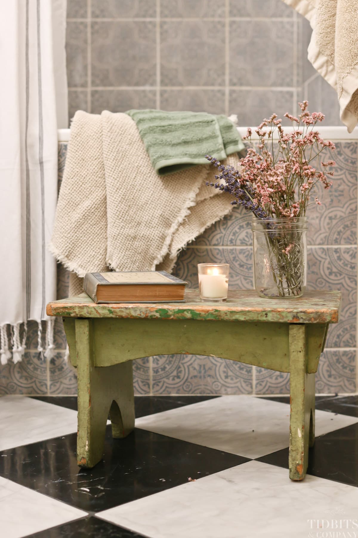 A green wooden bench holds a book, candle and flowers in a small split bathroom