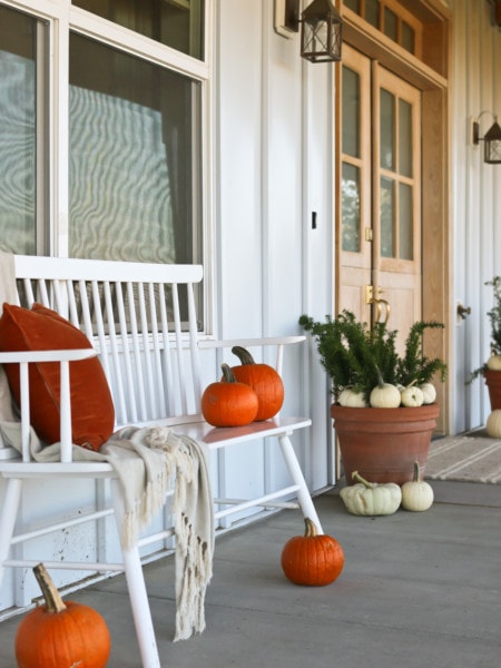 front porch decorated for fall with white and orange pumpkins plus a bench with pillow and blanket