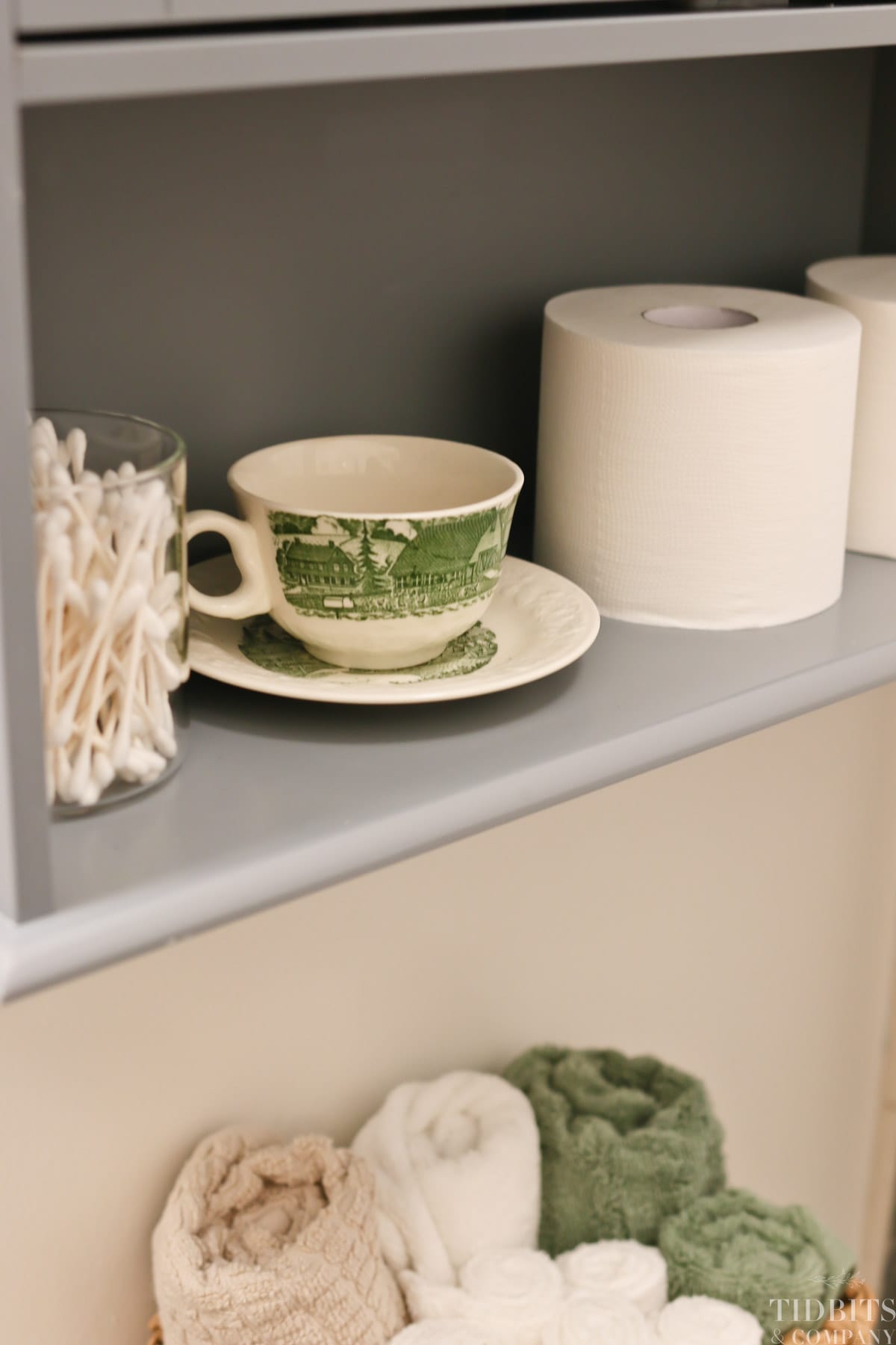 A tea cup, toilet paper and q tips on a bathroom shelf